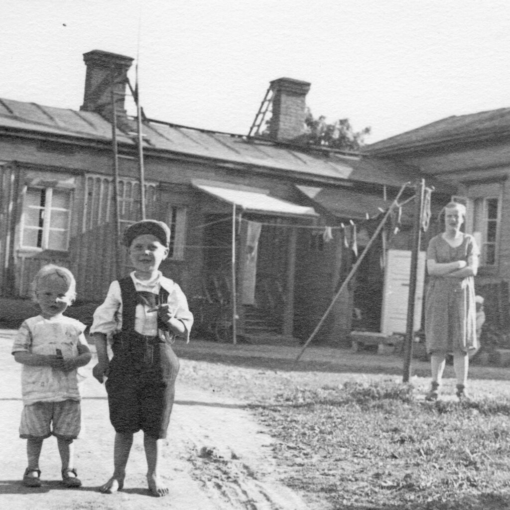 Mustavalkoisessa valokuvassa kaksi pientä lasta kuvan etualalla, takana puutalo sekä nainen.